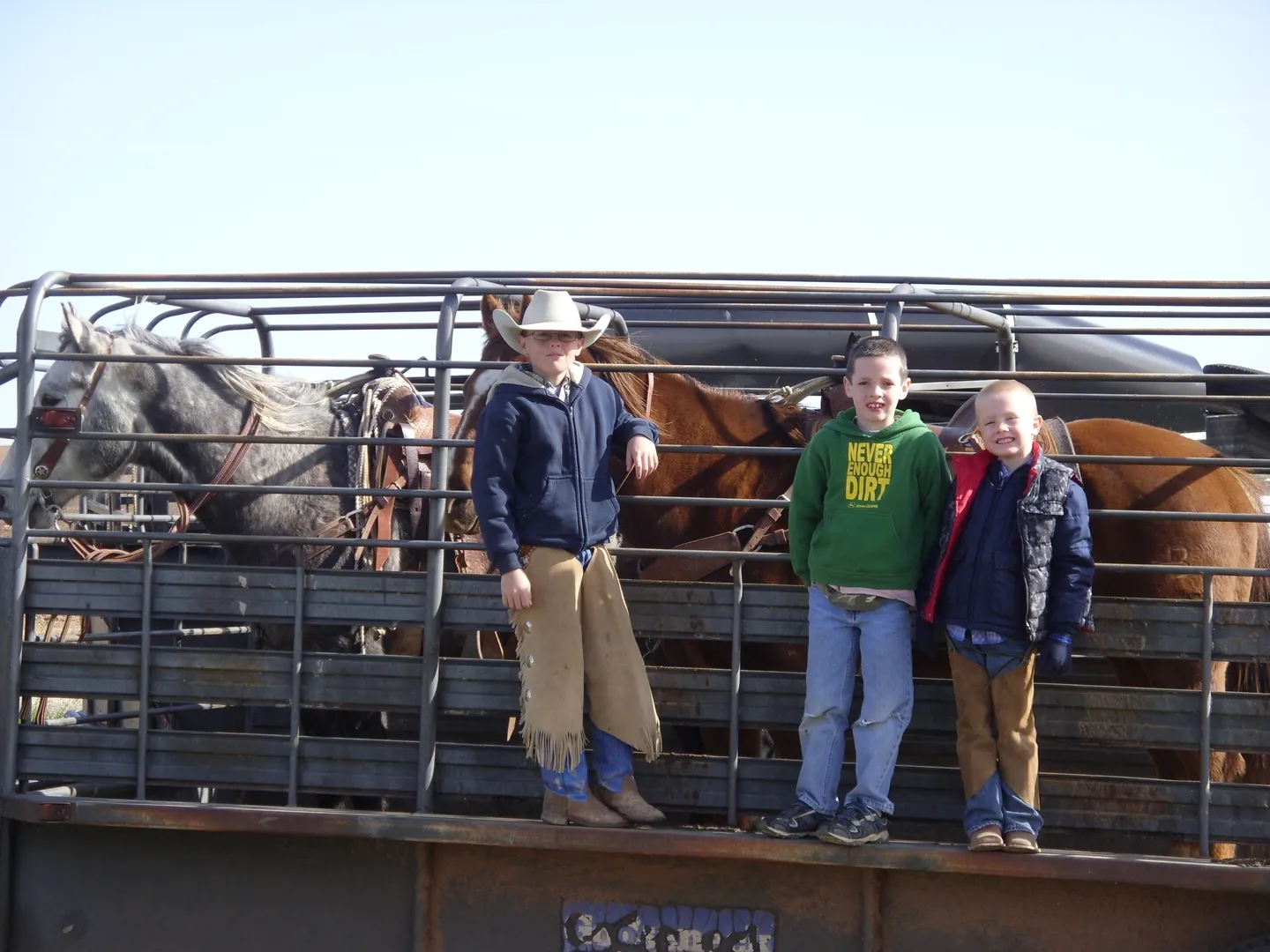 Three children standing on a truck bed with horses behind them.